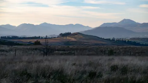 Lisa Birch A beautiful image of Scotland shows a wide, open grassland stretching across the foreground, dotted with sparse shrubs and muted earth tones.
Rolling hills rise gently toward layered mountain ridges fading into the distance.
Soft daylight and pale clouds create a calm, expansive atmosphere over the landscape