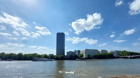 A tower block and some smaller buildings against a backdrop of blue sky and a few cumulus clouds with the River Thames in the foreground