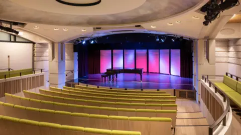 The auditorium at St John's College. It is small and modern with lime green seating and a large piano on the stage.