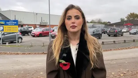 BBC Miss Hudson, who is a young blonde woman, stands outside the Excel carpark in question, wearing a brown coat with a poppy on the lapel