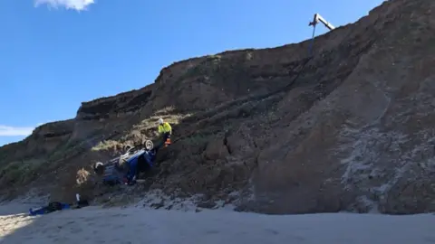 East Riding of Yorkshire Council A person dressed in yellow and orange hi-vis gear standing above an upturned blue Peugeot on a cliff. The sky is blue.
