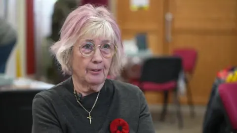 An elderly woman with grey and pink highlighted hair with glasses wearing a dark grey top with a chain and cross around her neck. 