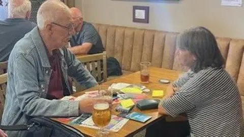 Karen Goddard Karen Goddard interviewing a dock worker. They are sitting opposite each other at a brown table. On the table are drinks, notes and a tape recorder