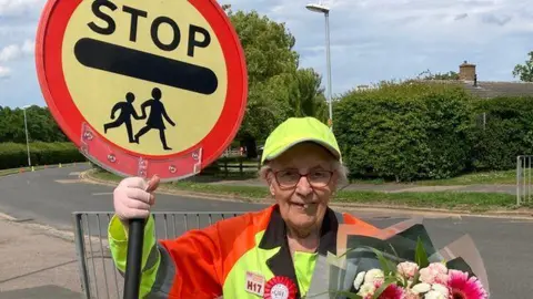 Carole in her uniform, on a roadside, holding a bunch of flowers in one hand and a "stop" sign in the other.