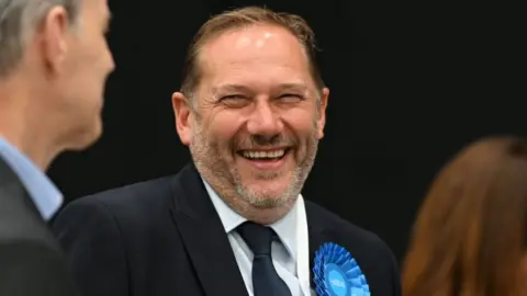 PA Media A antheral   with abbreviated  acheronian  hairsbreadth  laughs. He is wearing a concern  suit   and a bluish  rosette. 
