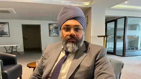 Clare Worden/BBC Harman Sond is wearing a grey suit and purple tie which matches his purple turban. He has a grey beard and wears glasses.