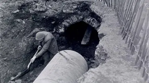 A black and white photo shows a worker digging in a trench beside a large cylindrical pipe, with a small tunnel opening in the exposed earth and a fence running along the edge.