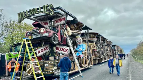 Andy Bennett A huge Somerset carnival cart sits at the front of a line of other carts on a main road. There's a ladder up on one side with repairs taking place. There's a few people looking at the carts.