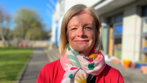 Nicola stands on a path beside a school building, wearing a red cardigan and a colourful patterned scarf. The background shows grass, trees, and outdoor play or garden items in bright sunlight.
