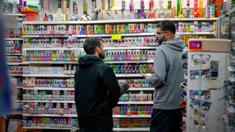 Shelves stacked with colourful vape products - e-cigarettes of all sizes and shapes. There are neon-coloured stickers saying 'No ID, No Sale' and others advertising offers and discounts. Sofy Saleem stands on the right, BBC reporter David Wallace Lockhart stands on the left.
