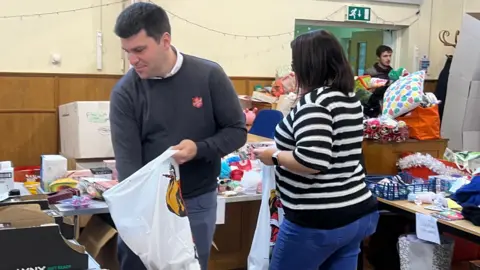 Two volunteers filling up hamper bags with items on tables in a hall