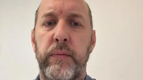 Rob Bolton Rob Bolton, a man with a beard and a blue shirt, sat in front of a pale wall