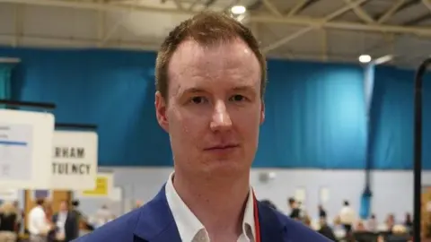 A head and shoulders image of the leader of Rotherham council. He has short ginger hair and is wearing a dark blue jacket with a white shirt.