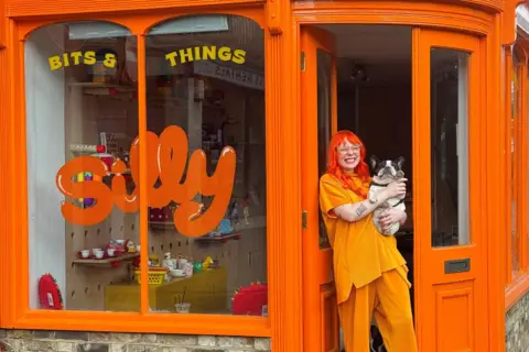 Sheri Scott Sherri Scott standing outside her shop, called Silly, which is painted orange. She is wearing orange clothes and has orange hair. She's holding a black and white dog - unfortunately not a ginger cat.