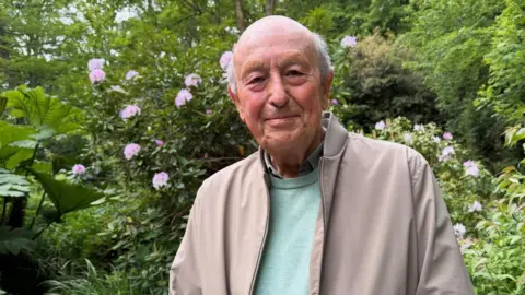 Tony Allchurch is wearing a brown jacket and green jumper. He is pictured in the Vallee Des Vaux peace garden. 