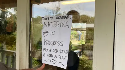A sign on a piece of a paper in a window saying "due to weather, watering still in progress. Please ask staff if need a plant."