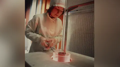 BT Group A female engineer wearing a white cloak and hat as well as clear gloves holds a pair of forceps to manoeuvre a piece of metal in a cylinder shape that is slightly glowing orange. 