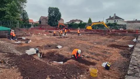 Cotswold Archaeology A group of archaeologists working at the site. There are excavators in the background and a member of staff in a high-vis jacket and helmet is working with hand tools in the foreground.