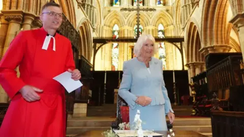 PA The Queen standing in front of a brightly lit Hexham Abbey altar. Beside her is a member of the clergy wearing glasses and dressed in red. There are flowers on a table.