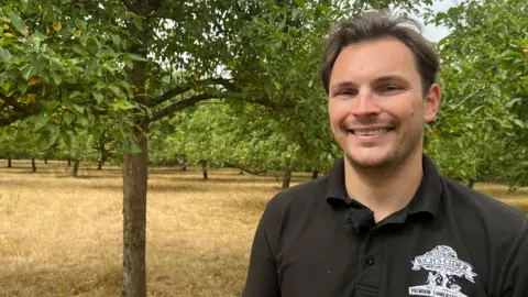 A young man wearing a black polo top smiles as he stands in an apple orchard. He has wavy but short dark brown hair
