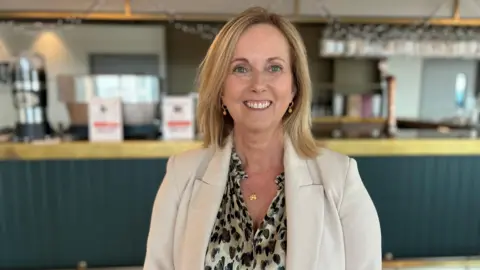 Geraldine Evans with short blonde hair, wearing a leopard print dress and white blazer.