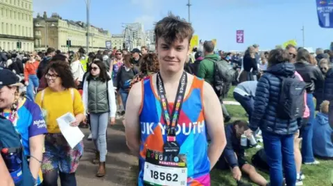 Billy Nunn, a young man wearing a bright blue and orange running vest, looks at the camera. He wears a Brighton Marathon finisher's medal around his neck. A crowd of people are standing and walking behind him. The sky behind him is blue.