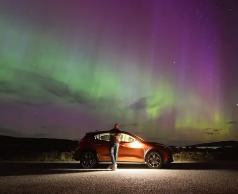 Gary Macleod The Northern Lights in the north coast of Scotland. There are green and purple ripples across the sky. Gary Macleod stands in front of his car under the lights.