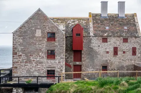 John O Groats Mill Trust Large stone three storey building with rusty red coloured window frames and shutters. There is a walkway with railings around the outside of the building