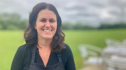 DJ McLaren/BBC A woman is smiling at the camera. She has dark brown hair and she is wearing a black top and a black apron