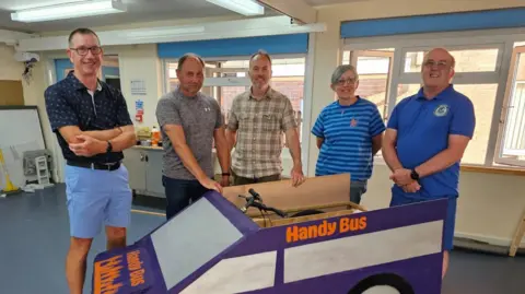 A group of five people pose for a picture. They are standing in front of a soapbox style vehicle which has "handy bus" printed on the side. 