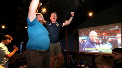 Two Scotland fans celebrate their team's win in a pub. A large TV screen in the background shows Scotland manager Steve Clarke. 
