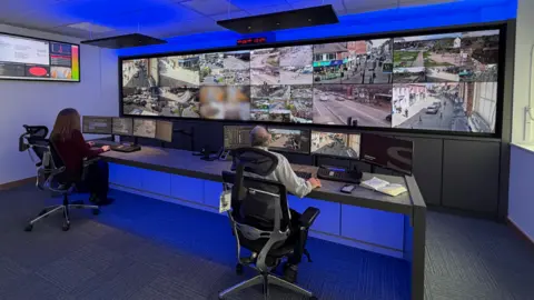 Two people sat at a desk looking at multiple screens with a blue light illuminating the desk.