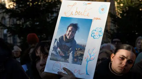 A person holds a photograph of Olivier Grondeau, showing him smiling, at a gathering in support of those arrested in Iran. 