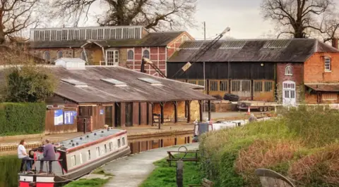 A collection of large buildings, some brick, some wood, with a canal leading to them and a narrowboat in the foreground with grass growing on the near side of the canal