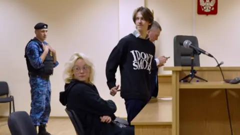 Diana sits in court while Alexander stands. A man in a blue combat uniform and a flak jacket looks on.