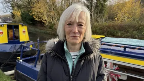 A woman with white hair in a dark coat stands in front of canal boats on the tow path, smiling at the camera