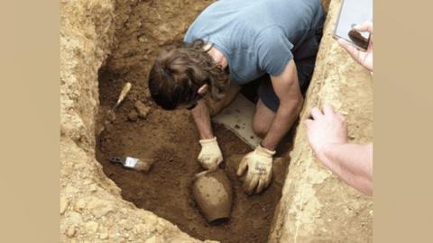 A student at the bottom of a deep hole in the ground uncovering a Roman pot from the mud