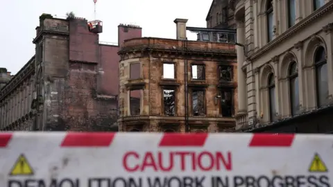PA Media Shell of a fire damaged Victorian building. A red and white barrier warning that demolition is in progress is in the foreground. 
