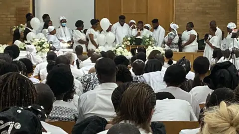 A large group of people dressed predominantly in white are gathered in a spacious hall. The audience is seated on wooden chairs, facing a stage at the front where individuals, also in white, are standing or seated around floral arrangements. The hall has brick walls, high ceilings, and large windows letting in natural light.