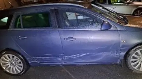 PSNI A blue car with damage along the door. It's parked at night and a silver car can be seen in the background. 