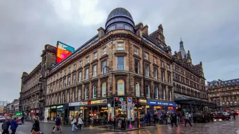 Getty Images Union Corner before the fire. The Victorian building has a domed roof on the corner and shops along the ground floor. People walk on the pavement in front of the buildings.