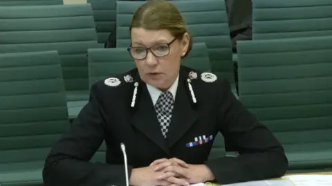 Parliament Judi Heaton is sitting behind a desk with her hands clasped together. She is wearing a traditional police uniform with her silver coloured badges clearly visible against her black jacket. She is wearing glasses and her brown coloured hair is being worn up.