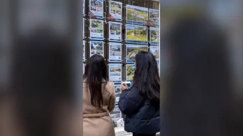 Two women with dark hair look at the window of an estate agent.