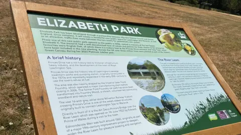 A wooden sign telling the history of the creation of Elizabeth park. It shows an image of the Queen Elizabeth and has text about the history of the area.