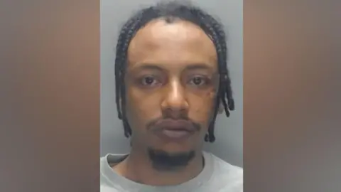 Mugshot of Mohammed. He has black dreadlocks, a thin black moustache and wispy black beard on his chin. 