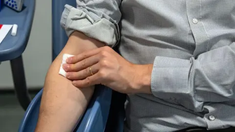 A man holds a cotton pad on his arm after blood has just been extracted from it using a syringe. He is in a hospital. 
