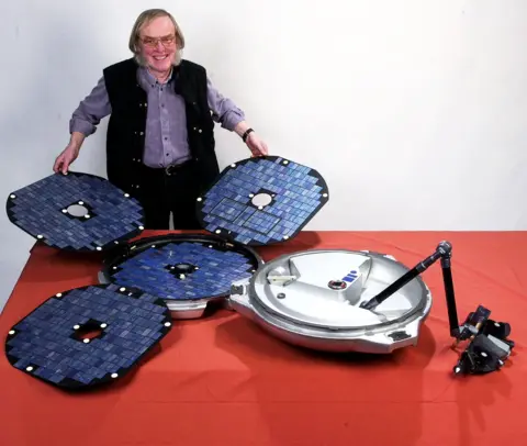 An older man with large sideburns is holding parts of a Mars lander, specifically solar panels
