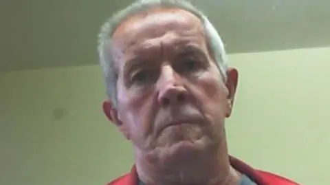 A low quality photo of Richard Scatchard. He has short white hair and no facial hair. He is pictured frowning and looking at the camera.