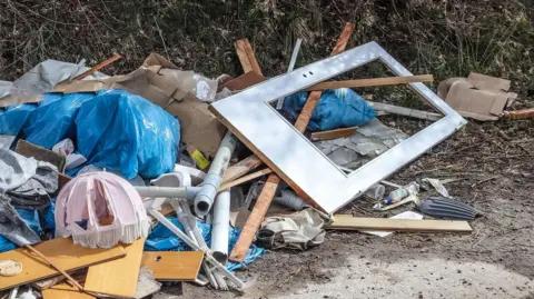 Generic fly-tipping at the side of a road, including a ripped lampshade and other rubbish.