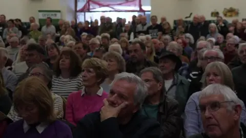 A wide shot of lots of people sitting inside a hall and listening to a presentation.